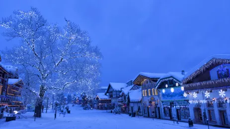 Snow covering a small town. A tree with twinkling Christmas lights and shop frontages decorated for Christmas