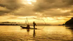 A fisherman in a boat throwing a net on the lake during sunset