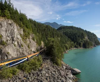 Aerial shot of dark blue and orange train surrounded by steep, rocky-mountains, tall pine trees and overlooking the ocean